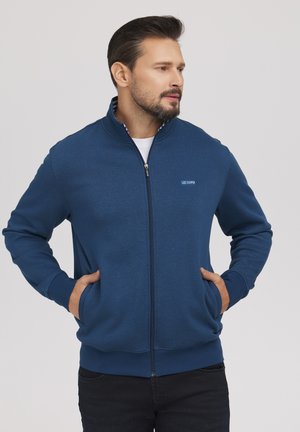 Mężczyzna z brodą, ubrany w niebieską kurtkę na zamek, z rękami w kieszeniach, patrzący w prawo na szarym tle.