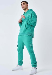 Turkis hoodie med en kengurulomme og matchende cargo-joggers med sidelommer. Båret med hvide sneakers for et sammenhængende look.