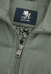 Helle graue Lederjacke mit einem silbernen Reißverschluss. Innen zeigt sich ein gemustertes Gewebe in dunklen Tönen. Ein Etikett mit einem Löwenlogo ist sichtbar.