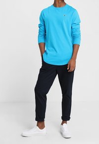Light blue long-sleeve shirt, black tapered pants, and white sneakers. The shirt features a round neckline and subtle logo detail on the chest.