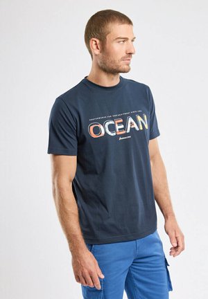 Mann mit kurzem Haar, der ein marineblaues "OCEAN"-T-Shirt und blaue Cargo-Shorts trägt, steht und schaut nach rechts vor einem schlichten Hintergrund.