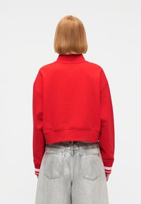 Άτομο με ίσια, ξανθά μαλλιά μέχρι τους ώμους, φοράει κόκκινο κοντό φούτερ και φαρδιά ανοιχτό γκρι τζιν, γυρισμένο προς τα πίσω σε απλό φόντο.