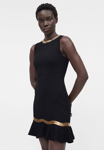 Γυναίκα που φοράει μαύρο αμάνικο φόρεμα με χρυσή επένδυση στη λαιμόκοψη και στο τελείωμα, στέκεται με τα χέρια πίσω από την πλάτη σε λευκό φόντο.