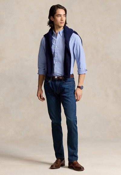 Mees, kes kannab sinist ruudulist särki, mille varrukad on üles keritud, tumedaid teksaseid, pruuni vööd ja kingi, sinist kampsunit õlgade peal, vaadates paremale.