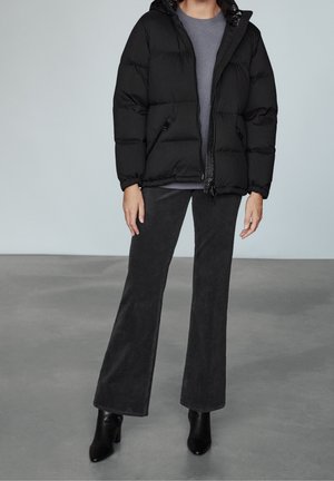 Person iført en sort polstret jakke, grå sweater, mørke udvidede bukser og sorte ankelstøvler med hæl, stående på en grå gulv.