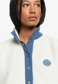 Weiße Fleecejacke mit einem blauen Druckknopfkragen und Logo. Verfügt über eine strukturierte Oberfläche und kontrastierende blaue Stoffakzente.