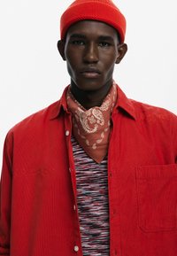 Rød kordfløjlsskjorte med en brystlomme, lagd over en stribet sweater. Et brunt bandana med hvidt paisley-mønster bæres om halsen.