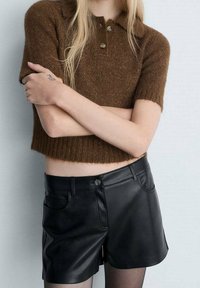 Top de polo de punto corto en color marrón con mangas cortas y tres botones, combinado con pantalones cortos de imitación cuero negros de cintura alta.