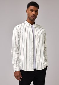 Valkoinen nappipaitaja, jossa on pystysuorat raidat sinisessä, punaisessa ja vihreässä. Pitkät hihat, rento kaulus ja pehmeä kangas. Puettuna valkoisen t-paidan päälle.