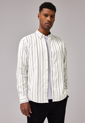 Camisa blanca de botones con rayas verticales en azul, rojo y verde. Mangas largas, cuello casual y tejido suave. Llevada sobre una camiseta blanca.