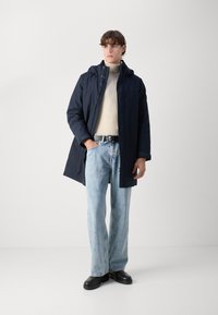 Marineblå vandtæt parka med hætte, båret over en beige sweater. Kombineret med lys blå brede jeans og sorte sko. Glat tekstur.