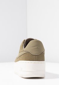 Zapatillas de ante verde oliva con una suela blanca gruesa, que presentan un acento cosido y un logo en la parte trasera. Diseño simple y limpio.