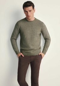 Ciemnooliwkowy sweter robiony na drutach z okrągłym dekoltem i prążkowanym wykończeniem, noszony z brązowymi spodniami, charakteryzującymi się teksturowanym, miękkim wykończeniem.