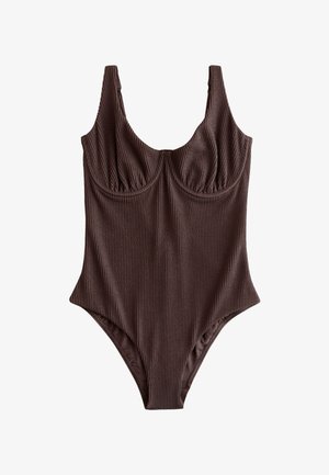 Frau in einem dunklen Einteiler steht mit den Händen im Haar, sieht auf den Ozean an einem sonnigen Strand mit Felsen und Wellen im Hintergrund.