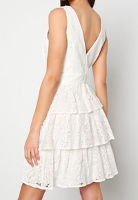 Abito bianco in pizzo con design a V sul retro, senza maniche e orli a strati con intricati motivi floreali. Texture morbida in tutto l'abito.