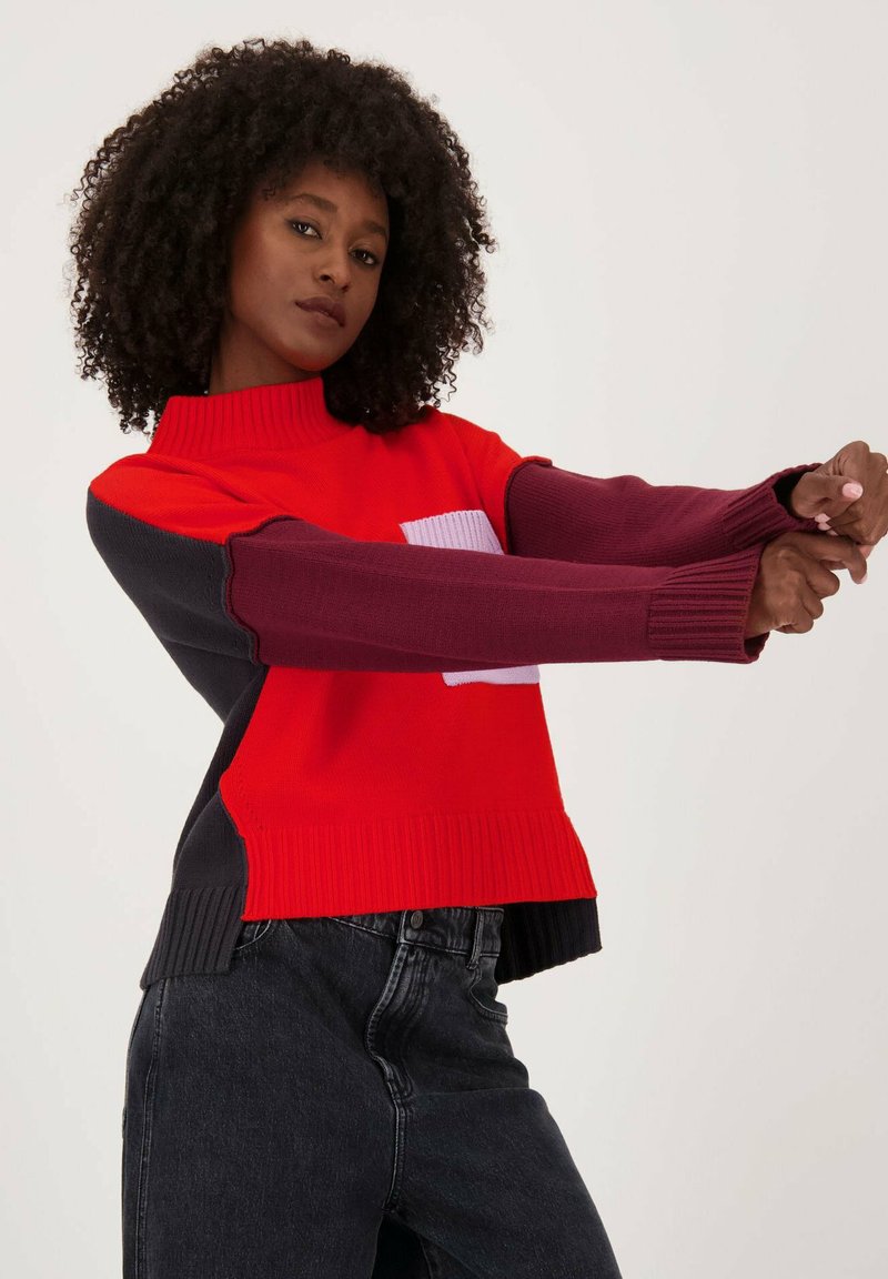 Rode en marineblauwe gebreide trui met geribbelde kraag en manchetten. Kenmerkend is het kleurblokontwerp met een roze accent. Het materiaal lijkt zacht en warm te zijn.