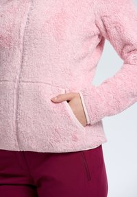 Chaqueta de forro polar de color rosa claro, suave y con cremallera, que presenta bolsillos laterales y una textura suave; combinada con pantalones de color burdeos oscuro.