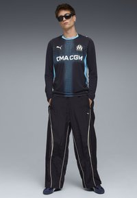 Czarny sportowy t-shirt z długim rękawem z niebieskimi akcentami i logo, w połączeniu z luźnymi czarnymi spodniami z białymi paskami i logo. Noszone okulary przeciwsłoneczne.