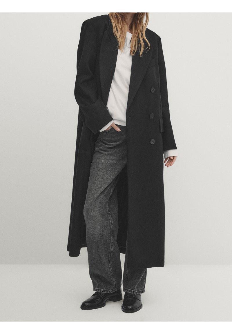 Lang sort frakke med skræddersyet design, dobbeltradet front og spidse klapper, båret over en hvid skjorte og grå jeans. Glat tekstur.