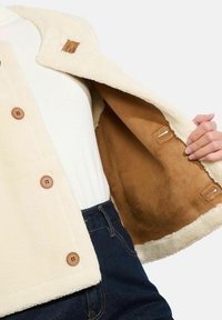 Krēmkrāsas pogām aprīkotais vilnas mētelis, atvērts, lai rādītu mīksto brūno iekšējo oderi, uzvilkts virs baltā krekliņa un tumši zilām džinsām.