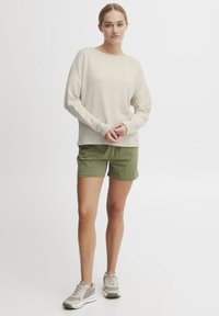 Pull en beige à texture avec un décolleté large, associé à un short vert olive. Le modèle porte des baskets grises avec des semelles blanches et des accents verts.