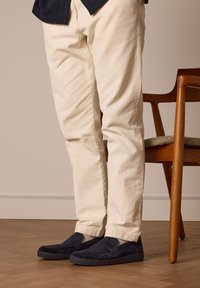Marineblauwe suède instappers met een gestructureerde donkere zool, gedragen met lichtbeige slim fit broek, tegen een houten stoel en een warme vloer.