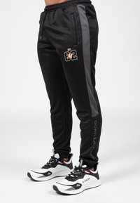 Zwarte sweatpants met grijze zijstrepen, elastische tailleband, zakken en gouden logo "EST. 82." Te combineren met zwart-witte sportschoenen.