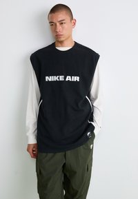 Sort ærmeløs Nike Air sweatshirt over en hvid langærmet skjorte, med fed hvid tekst og sideaksenter i en afslappet pasform.