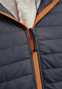 Grå polstret jakke med vandret syninger, orange lynlås og sort lynlås træk, over en lysegrå sweater med et mønstret krave.