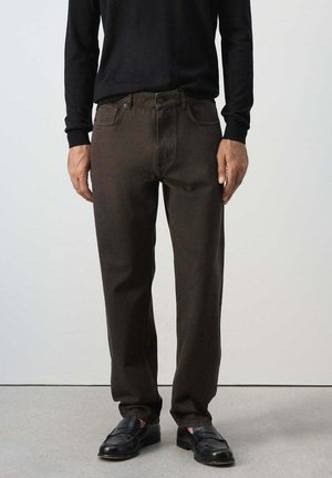 Mann trägt dunkelbraune gerade geschnittene Hose, schwarzes langärmliges Hemd und schwarze Leder-Loafer, steht vor einem einfarbigen Hintergrund.