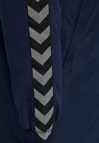 Mörkblå jacka med texturerad tyg, kontrasterande svart och vit chevrondetalj längs ärmen, med samlade muddar.