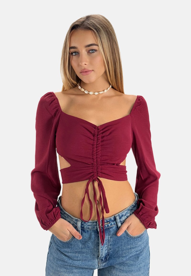 Bordeauxfarbener Cropped-Top mit langen Puffärmeln, ausgestattet mit Raffung an der Vorderseite und seitlichen Ausschnitten, kombiniert mit hoch taillierten blauen Jeans.