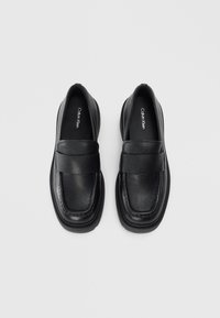 Schwarze Loafer aus strukturiertem Leder mit einem breiten Riemen über dem Spann. Sie verfügen über eine abgerundete Zehe und eine dicke Gummisohle.