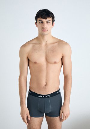 Boxer-briefs gris en mélange de coton, avec une ceinture noire arborant le logo "LACOSTE". Le modèle se tient de face, torse nu.