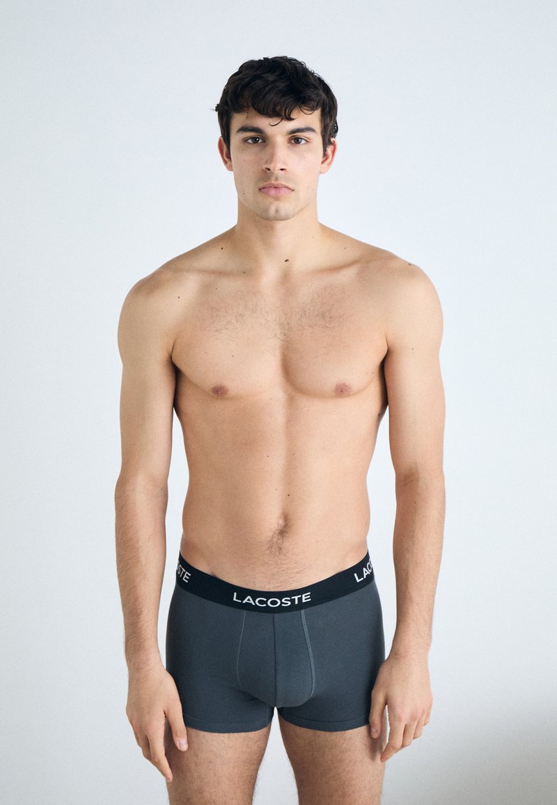 Graue Boxershorts aus Baumwollmischung mit einem schwarzen Bund, auf dem das "LACOSTE"-Logo zu sehen ist. Das Modell steht mit dem Gesicht nach vorne und ist oberkörperfrei.