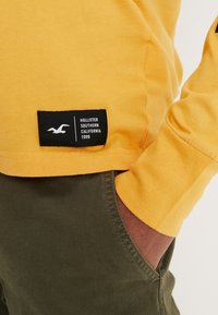 Gelbes langärmliges Hemd mit einem schwarzen Markenetikett am Saum, kombiniert mit olivgrünen Hosen. Der Stoff wirkt weich und lässig.