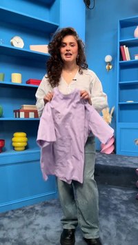 Kobieta z kręconymi włosami trzyma fioletową koszulę w niebieskim pokoju z półkami z książkami, zegarem i kolorowymi dekoracyjnymi przedmiotami.