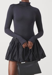 Body noir à manches longues associé à une jupe noire volumineuse, avec une texture lisse et un col roulé ajusté. Un petit sac à main noir complète la tenue.