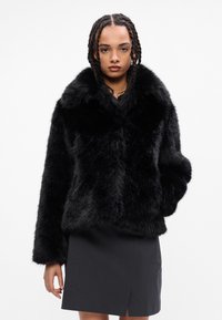 Casaco de pelúcia preto com um colarinho largo e mangas longas. A textura é fofa, oferecendo uma aparência suave. Usado sobre uma saia escura.