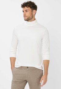 Maglietta bianca a maniche lunghe con collo a lupetto, tessuto morbido, design aderente, abbinata a pantaloni marroni a motivo, con polsini a costine e scollatura liscia.