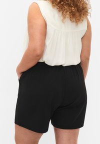 Person med lockigt hår som bär en ärmlös vit topp och svarta shorts, står med händerna i fickorna, vänd bort från kameran mot en vit bakgrund.