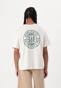Katoenen T-shirt in off-white met een groen cirkelvormig logo op de achterkant, met de teksten "WORLDWIDE" en "ESTABLISHED 1969." Relaxte snit.