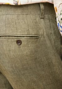 Oliwkowe lniane spodnie z boczną kieszenią i brązowym zapięciem na guzik. Tkanina ma teksturowany, lekko sztruksowy wygląd.