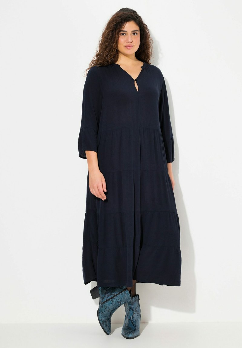 Navyblauwe, gelaagde maxi-jurk met een keyhole-halslijn, driekwartmouwen en een zachte textuur. Gecombineerd met blauwe enkellaarsjes met een slangenpatroon.