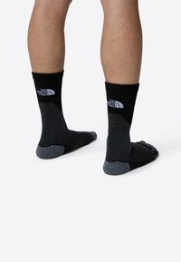 Calcetines negros y grises con una suela acolchada, que presentan un logo en la parte superior. Están hechos de una tela texturizada, con una altura a media pantorrilla y un diseño con separación de los dedos.