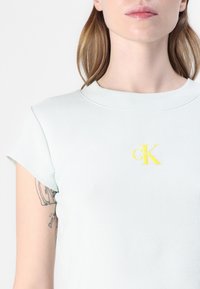 Světle modré tričko s žebrováním, kulatým výstřihem a krátkými rukávy. Významné žluté logo "CK" na přední straně blízko límce.