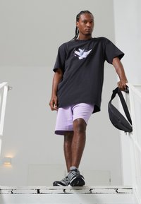 Camiseta negra de gran tamaño con gráfico de ave blanca, combinada con pantalones cortos morados claros. Zapatos grises y mochila negra de bandolera. Entorno en una escalera interior.