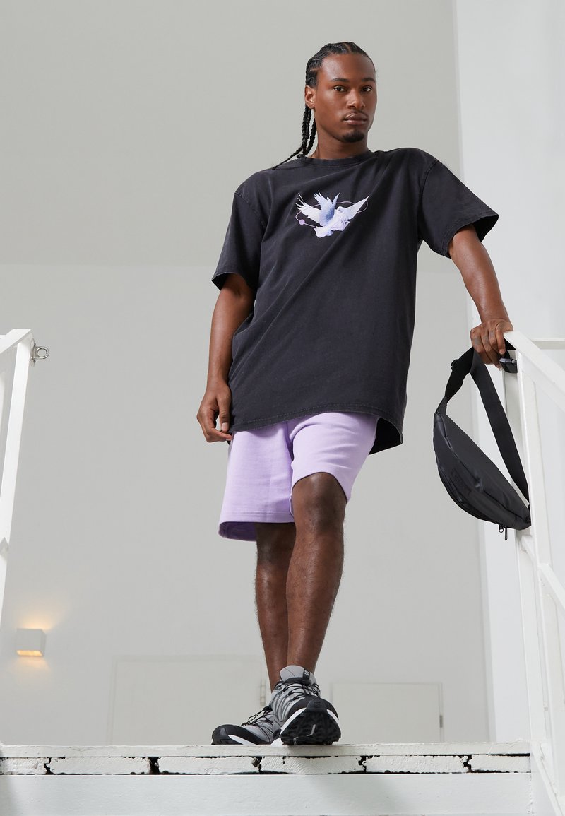 Camiseta negra de gran tamaño con gráfico de ave blanca, combinada con pantalones cortos morados claros. Zapatos grises y mochila negra de bandolera. Entorno en una escalera interior.
