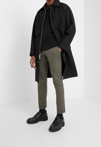 Czarny długi płaszcz, czarna bluza, oliwkowe slim-fit spodnie, czarne buty, z teksturowanym materiałem i detalem zamka z przodu.