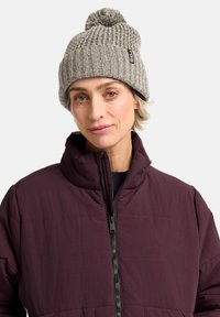 Beanie grigia lavorata a maglia con texture a coste e pom-pom sulla parte superiore, abbinato a una giacca imbottita bordeaux con chiusura a zip e colletto alto.